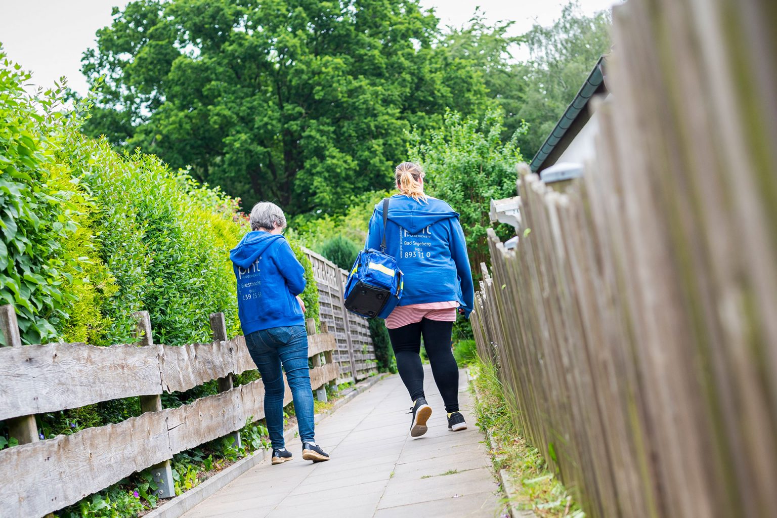 Zwei Menschen in blauen Jacken gehen mit Taschen in der Hand einen gepflasterten Weg zwischen Holzzäunen und Grünflächen entlang und entfernen sich von der Kamera.