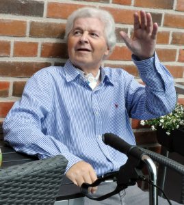 Ein älterer Mann mit grauem Haar sitzt an einem Tisch neben einer Backsteinmauer und winkt zur Begrüßung. Vor ihm ist der Lenker eines Rollators zu sehen.