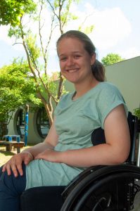Eine lächelnde junge Frau im Rollstuhl im Freien, trägt ein hellgrünes T-Shirt, mit Bäumen und einem Gebäude im Hintergrund.