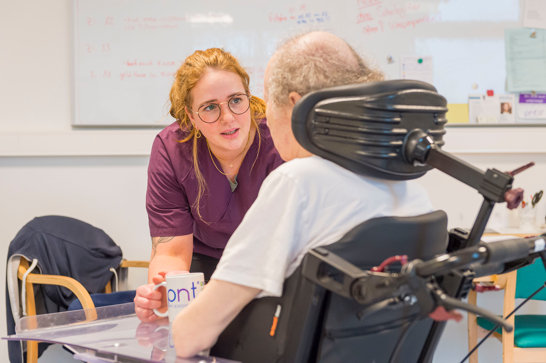 Eine Pflegekraft im lila Kittel spricht mit einer älteren Person im Rollstuhl und hält einen Becher in der Hand. Sie befinden sich in einem hellen Raum mit einer weißen Tafel im Hintergrund.