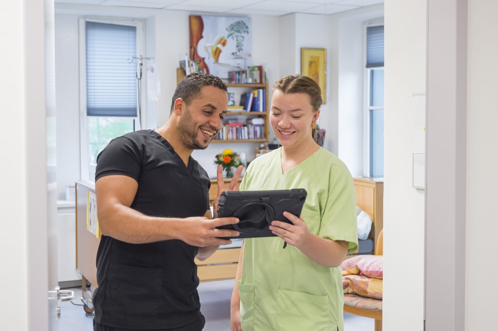 Zwei medizinische Fachkräfte stehen lächelnd in einem Behandlungszimmer und schauen gemeinsam auf ein Tablet.